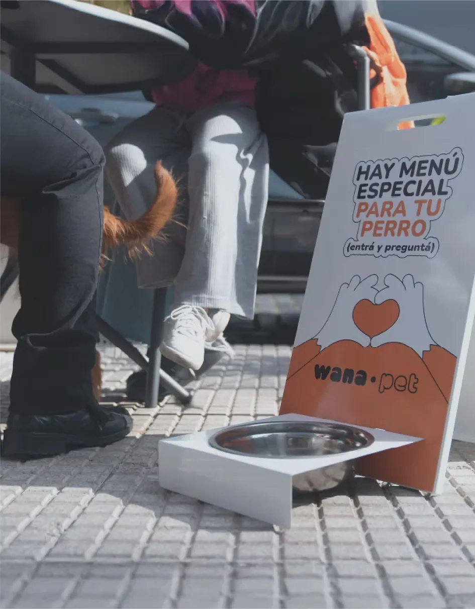 Mesa de café en la vereda con un perro y el cartel de Wana: hay menú especial para tu perro